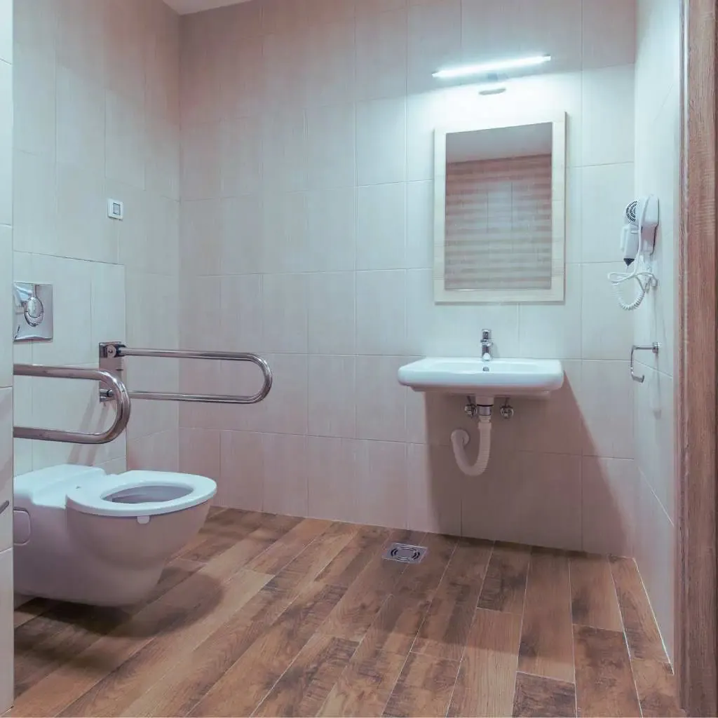 Accessible office bathroom showing wall-mounted toilet with side grab bars, sink with knee clearance, and wide turning space on a wood-textured floor compliant with California ADA design standards.