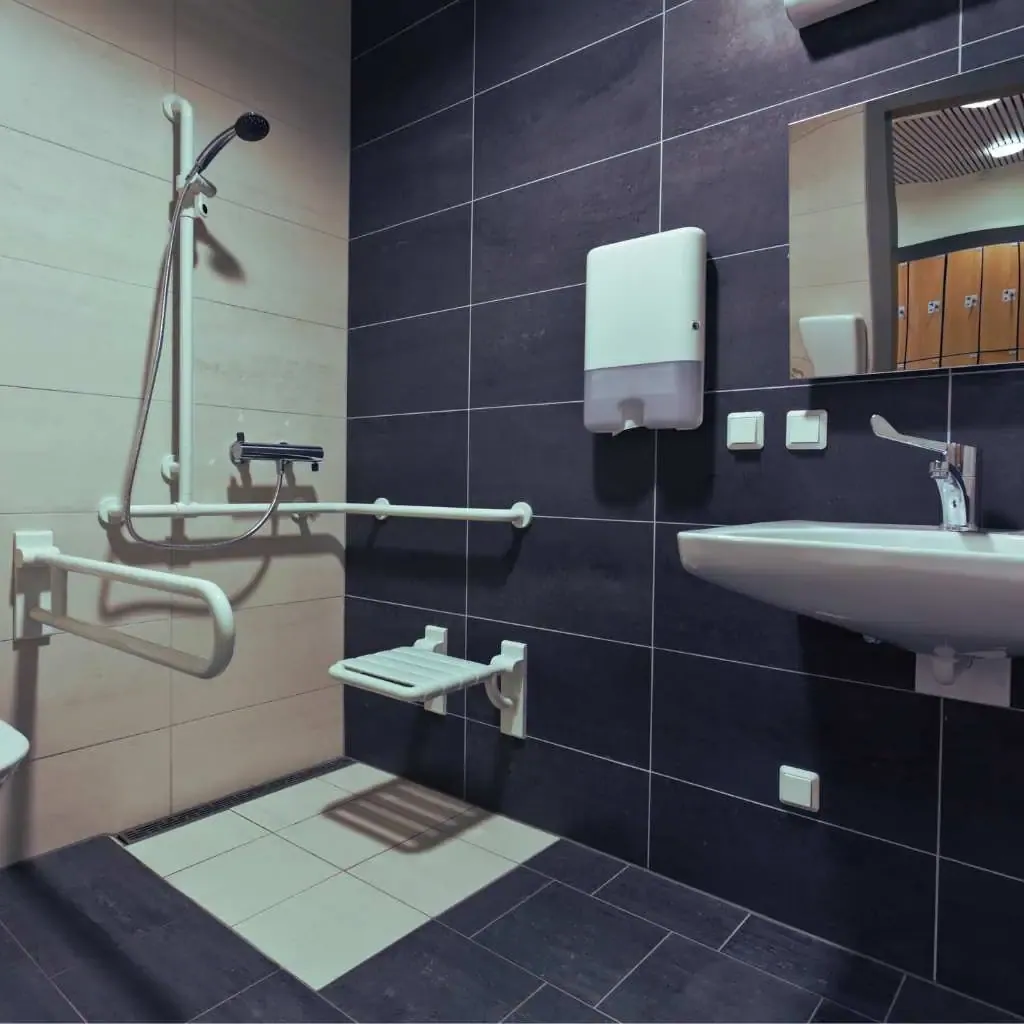 Accessible office bathroom with black tile walls, featuring a wall-mounted shower seat, grab bars, a floating sink, and ADA compliant fixtures