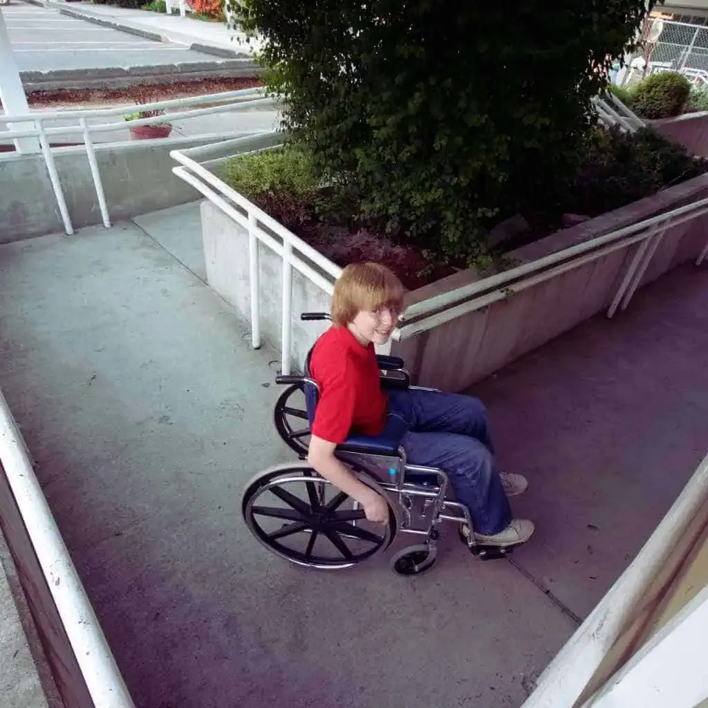 A smiling boy in a wheelchair uses a concrete ramp with handrails, demonstrating accessible design at a public facility in California.