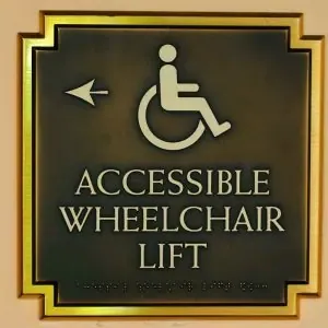 Bronze accessibility sign with wheelchair symbol and Braille, indicating the location of an accessible lift in a California building.