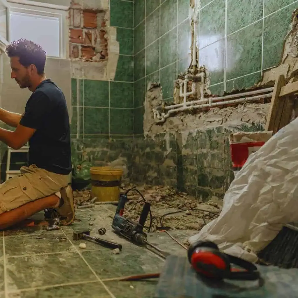 Contractor working inside a partially demolished bathroom during renovation, with exposed plumbing and tools on the floor, showing conditions before ADA and Title 24–compliant fixtures and clearances are installed.