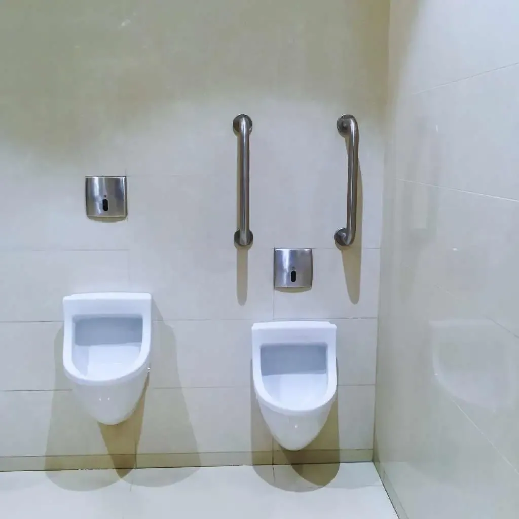Two wall-mounted urinals with vertical and horizontal grab bars installed to support users, showing an accessibility layout often required in California public restrooms.